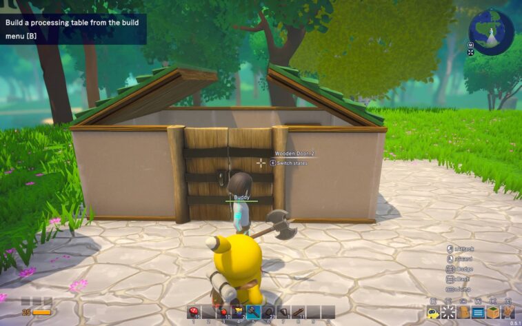 A sunny scene from Omega Crafter captures the buddy robot with a digital face screen and a hammer, in front of a partially constructed building with a wooden door. The player's character is seen from behind, interacting with the door, indicated by the floating text 'Wooden Door_2' and an action prompt 'E Switch states'. The tutorial overlay suggests building a processing table. The surrounding area is a well-trodden stone path bordered by lush green grass with small pink flowers, set against a backdrop of dense forest. Published on: LadiesGamers