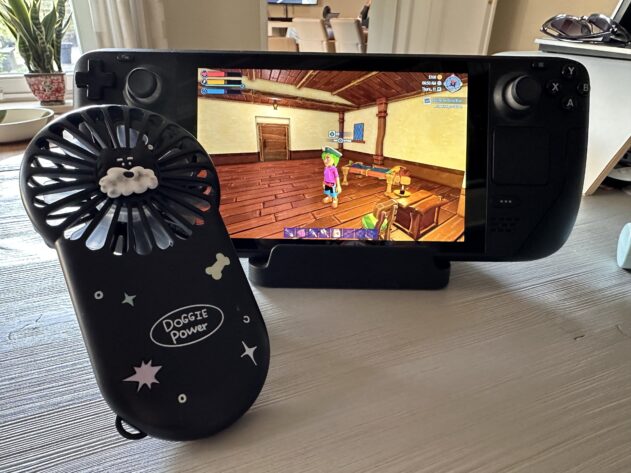 An image in a YvoCaro Plays article: A Steam Deck is displayed on a table, showing gameplay from the video game Moonstone Island. The game's screen features a character standing inside a wooden house with vibrant colors. In the foreground, there is a small handheld fan with a "Doggie Power" design, decorated with star and bone stickers. The room has a cozy atmosphere, with a plant on the windowsill and sunlight streaming in from outside. Published on: LadiesGamers.