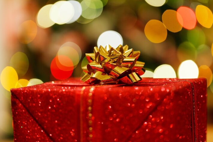  Favourite Christmas Gaming Memories An image showing a present wrapped in red wrapping paper and topped with a gold bow. In the background is bokeh created by the lights from a Christmas tree.