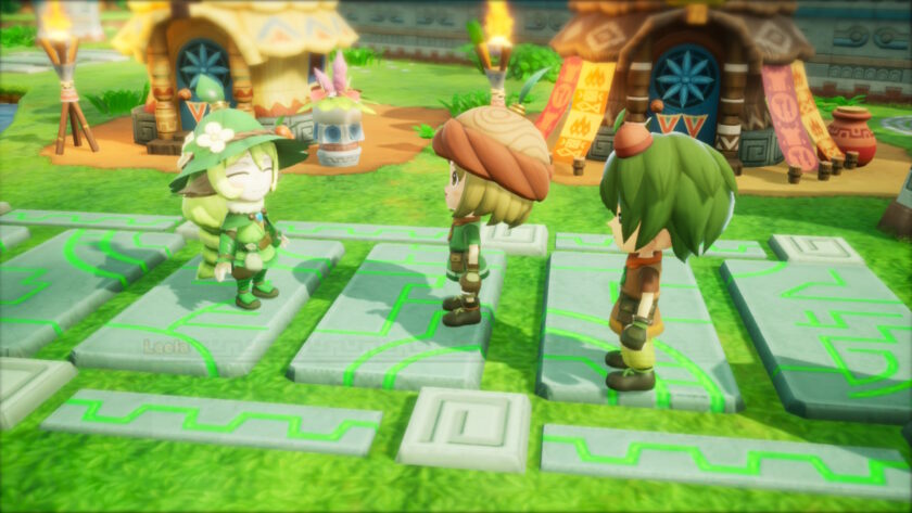 Three characters chat in Fantasy Life I outdoors in a vibrant village setting. One wears a leafy green outfit and flowered hat, smiling brightly, while the others stand attentively facing her. Round huts and totem-like torches decorate the background. Published on: LadiesGamers