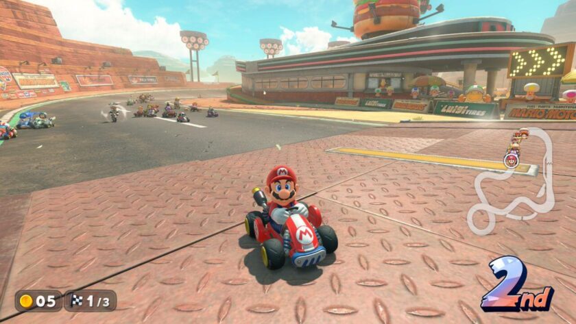 Mario drives a red go-kart past the starting line on a desert-themed race track in Mario Kart World, with a retro-style diner and giant burger structure in the background. Other racers follow behind him as he holds second place in the race, shown on the HUD. Published on: LadiesGamers.
