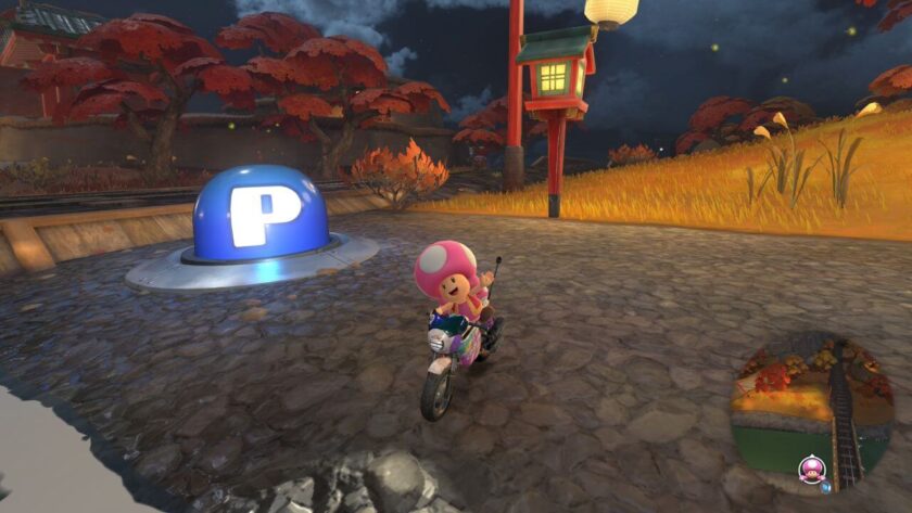 Toadette rides a motorcycle under a glowing lantern on a cobblestone path in an autumn-themed track in Mario Kart World. A large blue button with a white “P” is embedded in the ground nearby, surrounded by red-leaved trees and golden grass under a night sky. Published on: LadiesGamers.