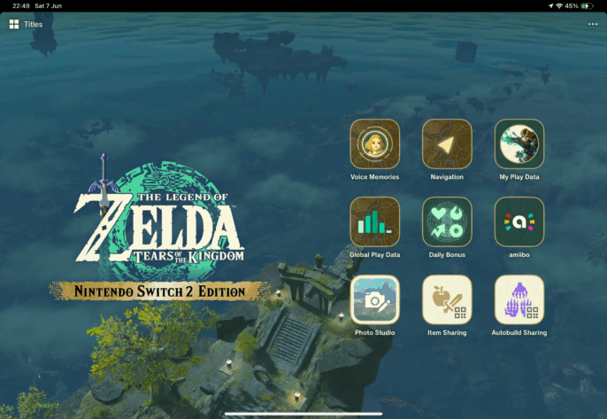 Main menu screen from The Legend of Zelda: Tears of the Kingdom - Nintendo Switch 2 Edition. The background features a misty aerial view of the floating islands and ruins of Hyrule. On the left is the game’s logo with a glowing green circular pattern behind the Master Sword. To the right are various menu icons: Voice Memories, Navigation, My Play Data, Global Play Data, Daily Bonus, amiibo, Photo Studio, Item Sharing, and Autobuild Sharing. Published on: LadiesGamers.